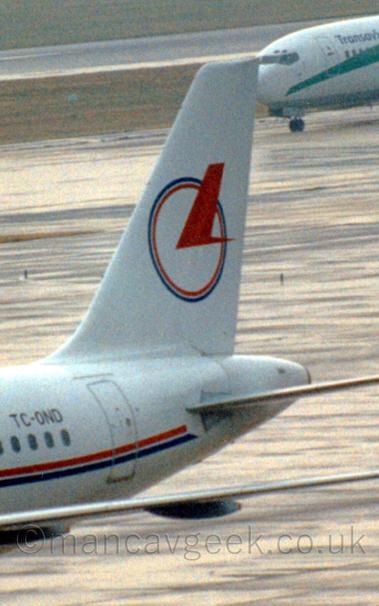 Close up of the tail of a white jet airliner, with a thin red and blue stripe on the fuselage, with  a circle intersected by a stylised red bird is on the tail.
In the background is the nose of another aeroplane with a green flash on the forward fuselage.