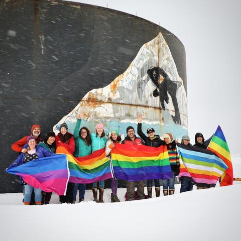 Twelve people stand with various rainbow flags in front of a large orca in front of an iceberg painting with faded paint.