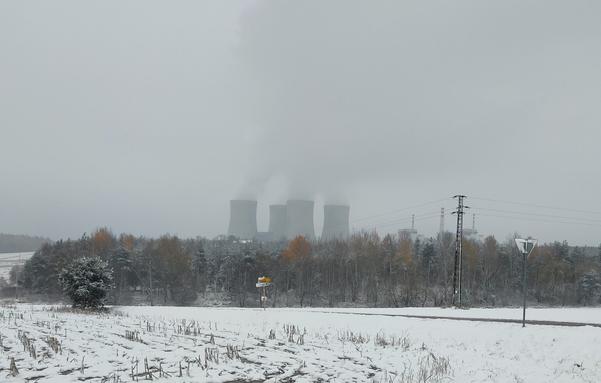 A last few trees still in their autumn leaves provide ghostly smudges of colour in a mostly drab grey landscape: bare, snow-dusted trees and a snow-covered field lie under a dull sky, all other colour leached by a thin mist. Behind the stand of trees, a smear of steam rises from the four vast grey cooling towers of Temelin nuclear power station.