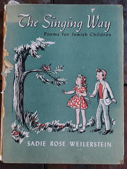 The cover of a large children's book, teal background with a tree on the left, a red bird on its branches, a boy and girl in black, red, and white looking at the bird from the right side of the cover. Title: "The Singing Way: Poems for Jewish Children," by Sadie Rose Weilerstein.