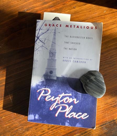 Paperback copy of Peyton Place by Grace Metalious, on a wood table. A gray pebble with faulted laminations rests on the cover.