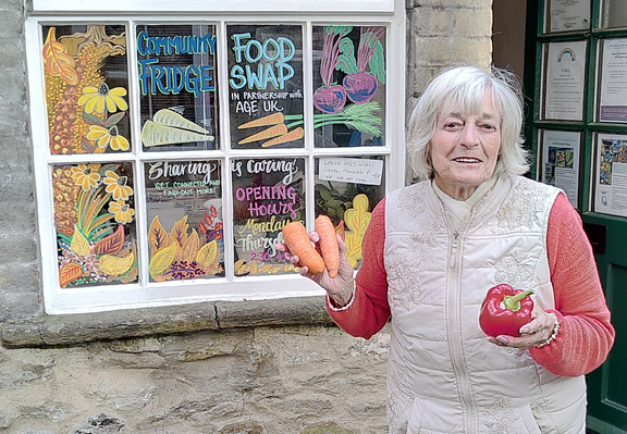 Picture of a woman with short white hair wearing a red and cream top whilst smiling and holding some carrots. Behind her is a window covered in colourful writing and images of fruit and vegetables with 'Community Fridge Food Swap' in large blue lettering