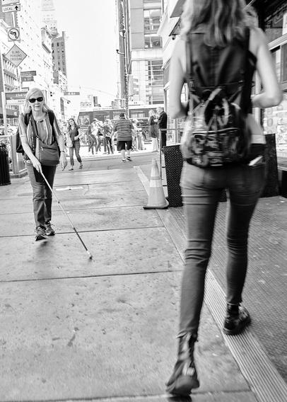 A blind person is traveling along a New York City sidewalk. A woman approaching from the other side is making an evasive maneuver to accommodate the blind woman's path of travel. Street bustle and a "Blind Persons Crossing" sign can be seen in the distance.