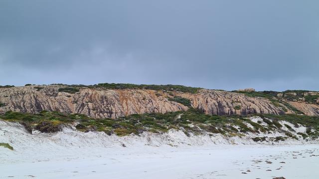 Dark and cloudy day, rocks and green shrubbery overlooking a white sand dune, on an Australian beach.