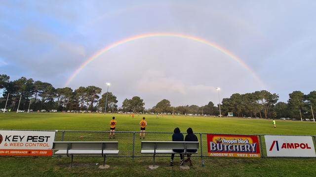 A blue sky almost completely covered by grey clouds. Australian rules football oval with signs lining the foreground advertising local business sponsors. Two players in black and gold wait to sub on while a game is underway. There is a complete rainbow in background, disappearing into the trees lining the far side of the oval. Two spectators sit huddled on bench in front of fence, in black hoodies with the hoods up against the cold wind.