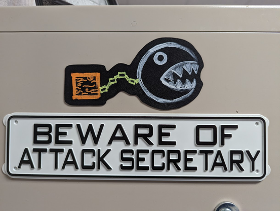 Two signs on a plain tan cabinet. One is a cut-out decal of a hand-drawn Chain Chomp from the Mario line of video games. The other says in plain font, "Beware of Attack Secretary."