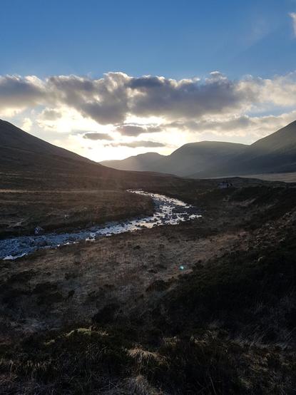 A photo of a Glen at sunset. Hills rise in silhouette on either side, a burn, shining silver in the low light, winds through the foreground and round behind one of them. The sun has just dipped behind a hill so rays are shining out.