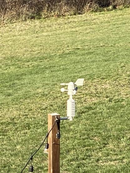 A weather station that measure wind speed and direction sits on a pole in a backyard.
