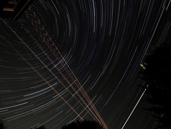 Night sky on London outskirts showing star trails around Polaris and crossed with plane vapour trails. Bright curved line of Jupiter bottom right.