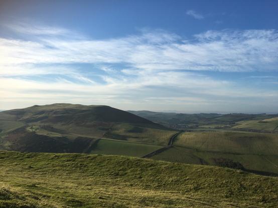 A landscape photograph of some rolling hills with a small village in the distance. The sky is light blue and there are whispy clouds. The scene is bathed in a warm sunny light.