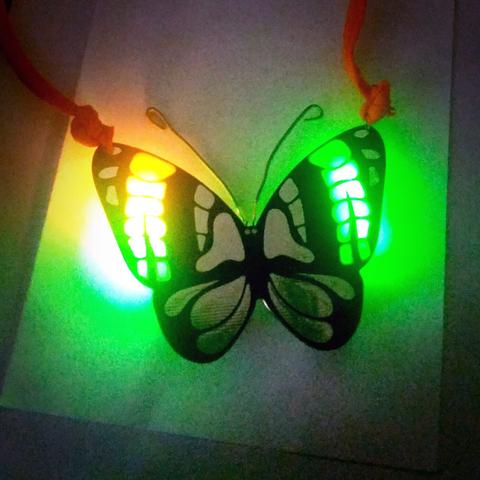 eine semitransparente Platine in Schmetterlingsform, die von hinten durch Farbwechsel-LEDs grün und gelb erhellt wird. An den Flügeln ist ein oranges Halsband festgeknüppert.