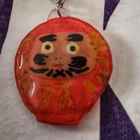 This daruma looks like a big round red ball, with a pink face and a confused expression. It must be confused to be involved in the ambition to create a daruma necklace. It is glossy and partially translucent.