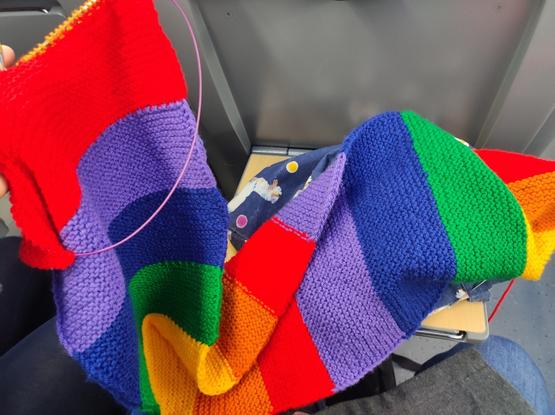 Langer Wollschal in Regenbogenfarben. Oben sind noch Stricknadeln im Werk. Das ganze auf ist auf einem kleinen Zugtischchen drapiert.