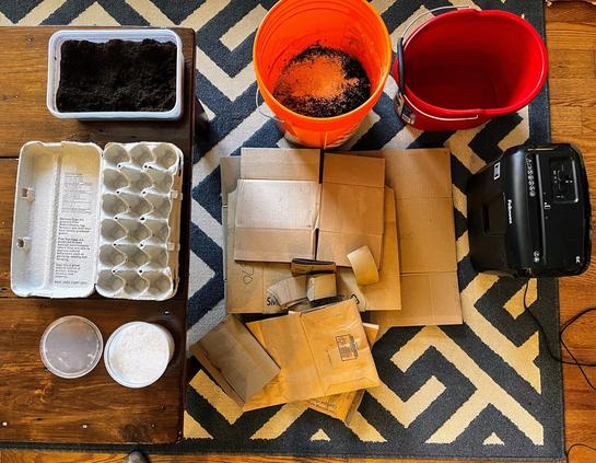 Mulch ingredients laid out: buckets, paper shredder, coffee grounds, egg cartons, ground eggshells, paper bags and cardboard.