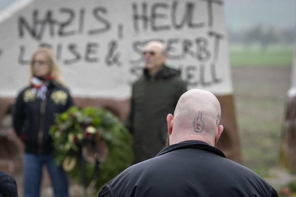 Ein Nazi-skinhead mit einem tätovierten Mittelfinger und dem Logo der Rechtsrockband "Lunikoff Verschwörung" auf dem Hinterkopf. Im Hintergrund das Mahnmahl "Feld des Jammers" auf dem mit schwarzer Farbe gesprüht steht "Nazis, heult leise und sterbt schnell".