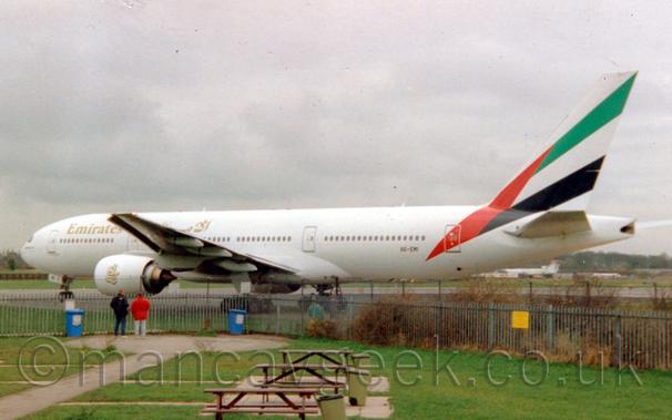Large white jet airliner with golden "Emirates" titles and a large UNited Arab emirates flag on the tail and rear fuselage.
In the foreground are two people watching it turn on to the runway, almost close enough to touch it.