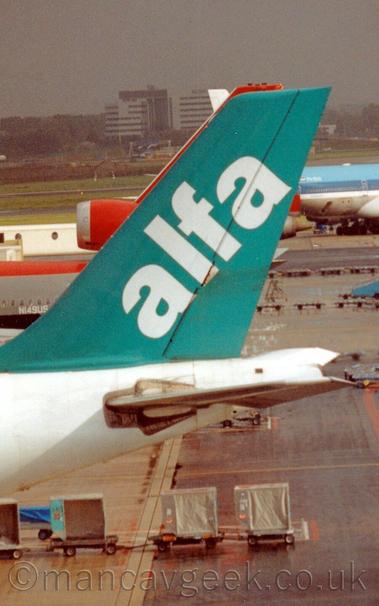 Closeup of the tail of a large white airliner with a mint green tail, with the word "alfa" written diagonally in lower-case white letters.
In the background is a busy airport scene, with a couple of planes parked or moving around.