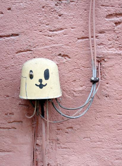Eine Verteilerdose an einer pinken Wand ist mit einem Gesicht bemalt und erinnert so an eine Qualle