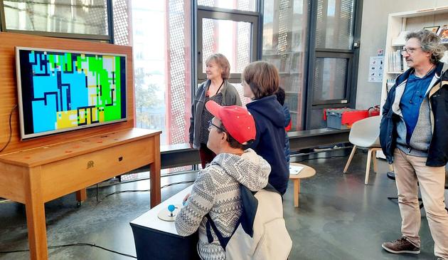 Two kids and and one adult play Dead Pixels, a game by Tatiana Vilela dos Santos Olivier Drouet while two adults, a man and a woman, watch them.
A big screen displays a colorful geometric graphic composition. Players hold colorful joysticks mounted on a big white board covered with painter's palettes.