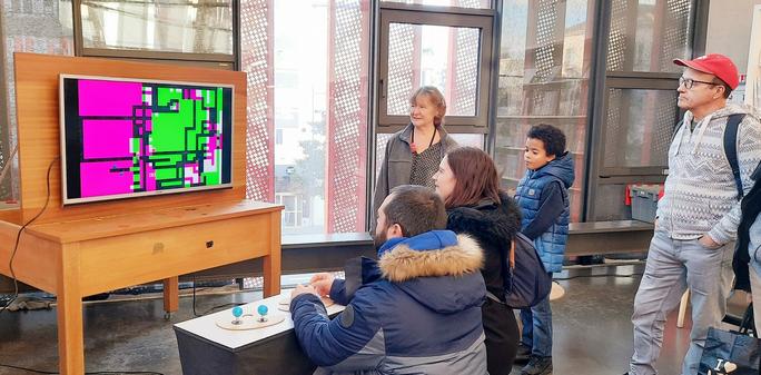 A couple plays Dead Pixels, a game by Tatiana Vilela dos Santos and Olivier Drouet, while a man, a woman and a kid watch them. 
A big screen displays a colorful geometric graphic composition. Players hold colorful joysticks mounted on a big white board covered with painter's palettes.