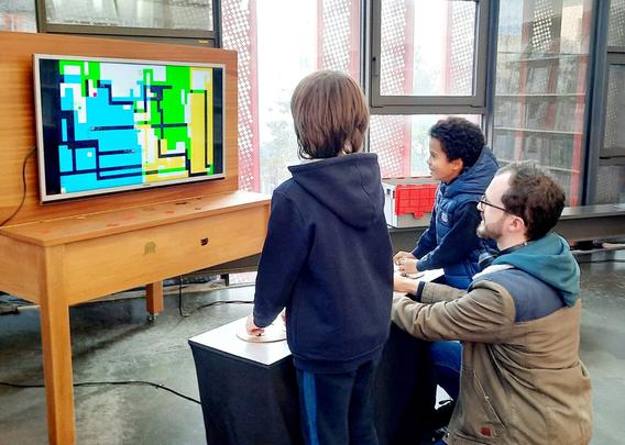 Two kids and a man play Dead Pixels, a game by Tatiana Vilela dos Santos and Olivier Drouet.
A big screen displays a colorful geometric graphic composition. Players hold colorful joysticks mounted on a big white board covered with painter's palettes.