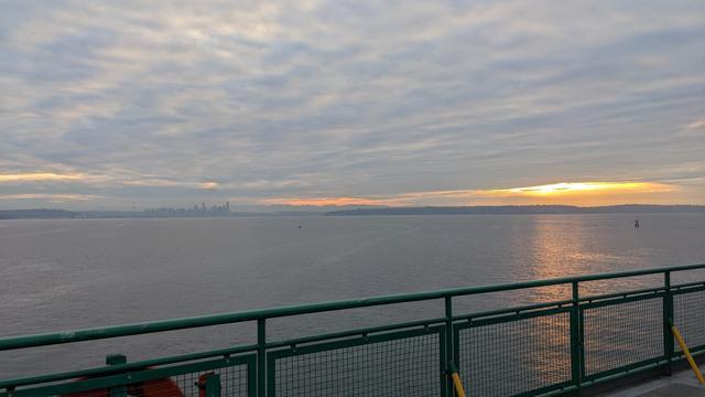 A small part of the sunrise glows orange and red, peaking out of a hole in the blanket of clouds. The colors reflect on the waters of Puget Sound, and the green railing from the top deck of the MV Tacoma is in the foreground.