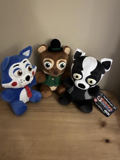 Three Five Night’s at Freddy’s series plushies set on a table next to eachother. From left to right, Candy the Cat, Popgoes the Weasel, and Blake the Badger.