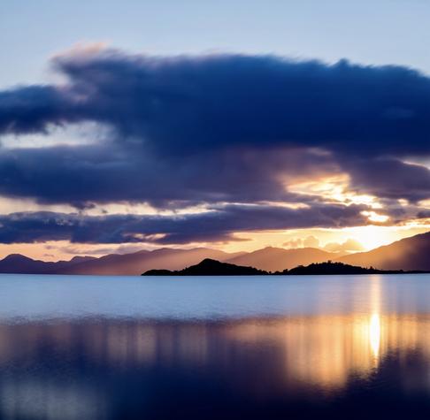Golden beams of light are cast across the calm waters of Loch Linnhe on the west coast of Scotland. The sun breaks through a layer of cloud before it sets behind the Morvern peninsula. An island sits in the loch, silhouetted against the sunlight.
