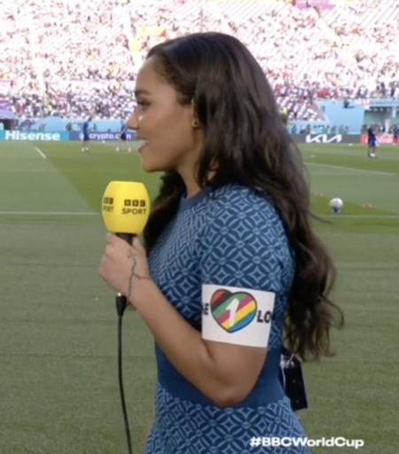 Presenter Alex Scott wearing the OneLove arm band holding a mic at side of the football pitch