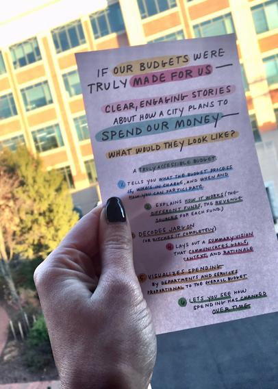 Photo of a hand holding a piece of paper. The page has part of an Ariel Aberg-Riger comic, that says:

“If our budgets were truly made for us—clear, engaging stories about how a city plans to spend our money—what would they look like? 

A truly accessible budget:

1. Tells you what the budget process is, who’s in charge, and when and how you can participate.

2. Explains how it works (the different funds, the revenue sources for each fund).

3. Decodes jargon (or ditches it completely).

4. Lays out a summary vision that communicates goals, vision, and rationale.

5. Visualizes spending by departments and services proportional to the overall budget.

6. Lets you see how spending has changed over time."