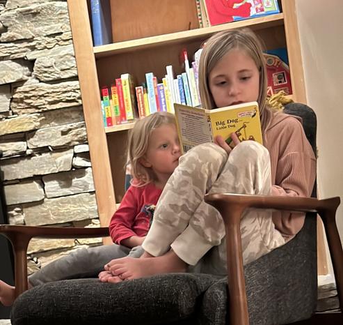 Blonde haired 10 year old girl is curled up with blind haired 3 year old girl in a mid century modern chair in front of a fireplace. There is a shelf of books behind them. The older girl is reading to the younger.