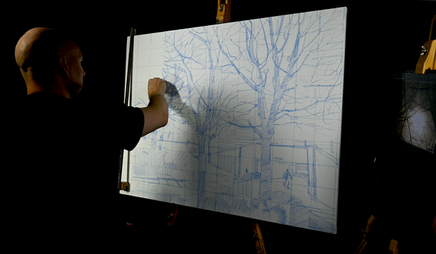 A large rectangular painting is in the early stages of being drawn in with blue pastel. Rough angular, marks fill the canvas over the top of a drawn in grid. Andy's hand can be seen drawing. the scene shows trees, pavement and a small drawn in man can be seen on the right side of the canvas.