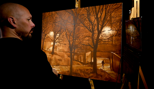 This photo shows an image of a cityscape painting. The view is of St Mary's church, here in Sheffield as seen from inside the nearby roundabout. The picture shows a church with a glow where the clock face should be. Silhouettes of trees fill the scene and a bank of grass leads up to the side of the road. A small man can be seen walking into an entrance to an underpass. This layer is an underpainting layer. All the forms have now been defined and it's very clear a night scene is shown. Everything has been painted in a singular warm , dark brown tone. Andy can be seen in profile working out what is left to be painted in.