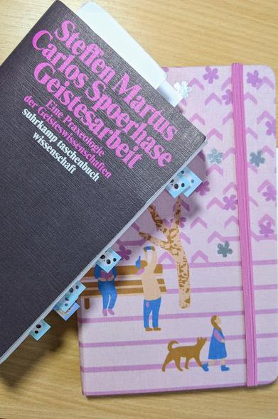 Suhrkamp Taschenbuch Wissenschaft "Geistesarbeit. Eine Praxeologie der Geisteswissenschaften" von Steffen Martus und Carlos Spoerhase liegt auf einem pinken Notizbuch, das zum pinken Schriftzug passt. Das Notizbuch hat eine japanische Illustration (Parkbank, Menschen, Kirschbaum) auf dem Cover und im Buch stecken Panda-Post-Its
