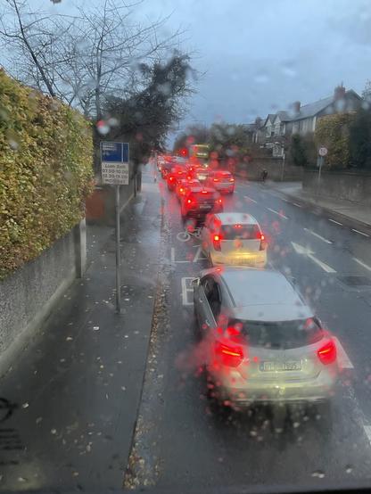 View out of the rain spattered front window of my bus showing a queue of about ten cars in the bus lane with brake lights glowing through the rain.