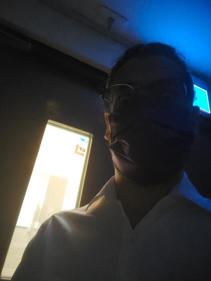 "A person in a dimly lit room, in front of a door with bright light shining through a window in the door. The person wears a facemask and glasses and has eyes widely opened, looking into the camera."