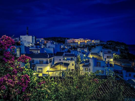 A colourful night shot of the village of Burgau in Portugal. White buildings and some pink Bougainvillea in the foreground.
