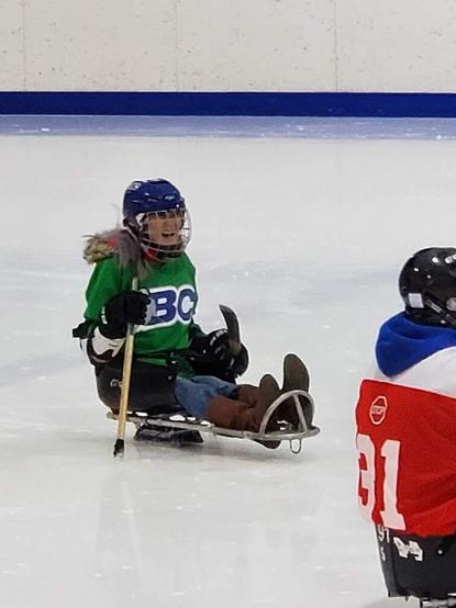 Me at sledge hockey practice, laughing with other players, holding my sledge hockey sticks, seated in my sledge skate.