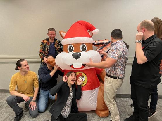 Group of seven men and a woman around an inflatable Buc-ee beaver, making funny faces or turned away as the beaver partially deflates