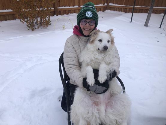 White and tan dog is laying in the lap of a woman with his stomach exposed. She is smiling and wearing winter clothing because there is a lot of snow on the ground.
