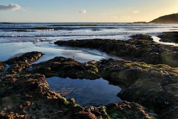 Pic taken low over rockpools reflecting blue sky with incoming waves and headland in the background.