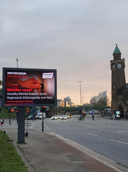 Hamburg, Landungsbrücken. In der Ferne die Elbphilharmonie. Eine breite Straße, wenig los. Neben dem Turm eine grelle Werbetafel. Auf dem Gehweg vorne eine zweite, riesige digitale Werbetafel mit dem Motiv "Hamburg dreht das. INFOSCREEN wir drehen das! Stecker raus! Standby-Betrieb braucht Strom. Ungenutzte Elektrogeräte vom Netz."