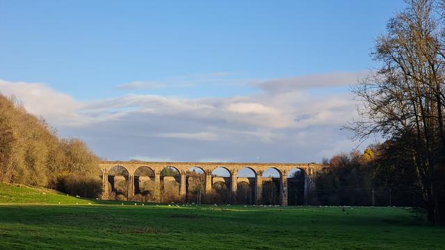 Chirk Viaduct and Aqueduct. The viaduct is higher than the Aqueduct. Built of stone locally quarried. In the foreground are green grass fields with sheep grazing. The trees on the left edge a road and the trees on the right edge the river. The sky is blue with low cloud on the horizon. The sun is shining and highlighting the Aqueduct casting a golden glow.