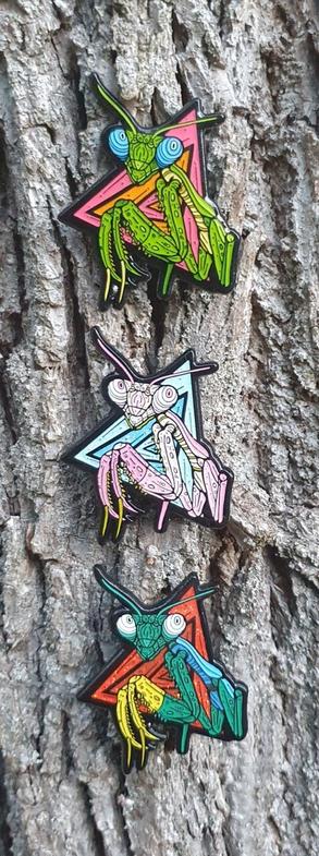 Three praying mantis enamel pins on tree bark.