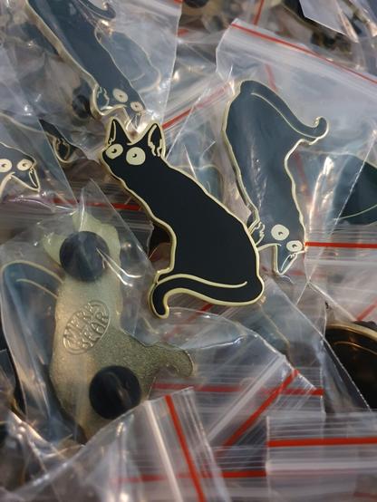 Black cat with a wide eyed stare enamel pin on a pile of other pins.