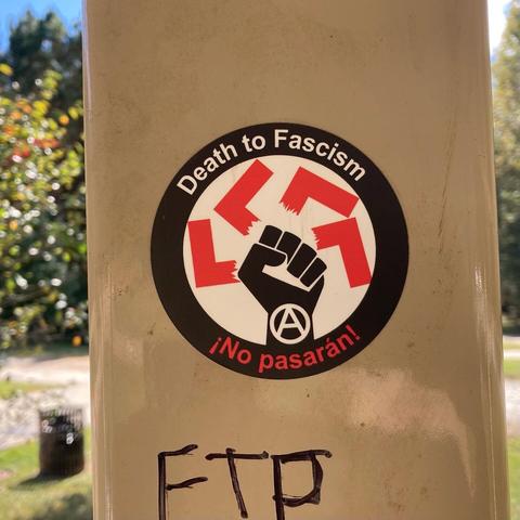 Red and black circular sticker with a drawing of a fist (with a circle A inside it) smashing a swastika, encircled by the words “Death to Fascism” and “No Pasarán.”
