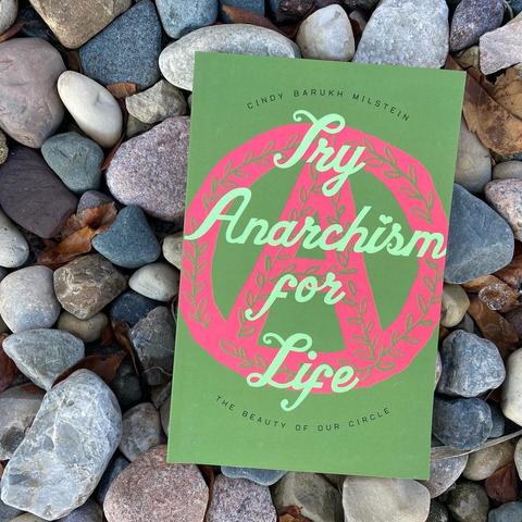 Book laid on a bed of gray-toned rocks; the book’s cover is two tones of green with the words “Try Anarchism for Life” overlaid on a pink drawing of a floral-patterned circle A.