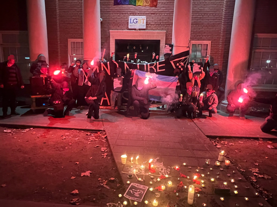 A picture of the front of a brick building with four massive pillars in front. The building is marked "Sacramento LGBT Center." Over two dozen people pose with various signs, road flares, pepper spray, candles and a bat behind a lit alter and in front of a hanging black banner. Many vigil goers throw up a power symbol or are wearing pink bandanas. Faces and shoes have been blurred.