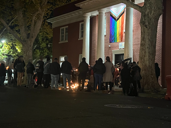 A giant, brick building with massive white pillars has a crowd of 50+ vigil goers with candles in front of an alter. A giant Queer Pride flag hands from the building above. Many vigil goers are in jackets, hoodies and various coats from the cold.