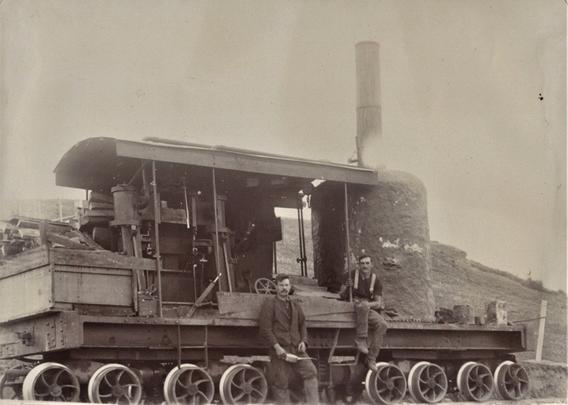 A black and white photo of a homemade woodfired boiler made to run the Clifden Limeworks plant back in the day when Jim Laurie set it up with some local lads. It became a large employer later and is still in operation today. This is an old homebuilt contraption with rail trolley wheels on a platform and an engine with a metal cover over it, a stack of wood to burn on the left and a large boiler with a chimney on the right. Two unknown men lean against the machine in work gear.